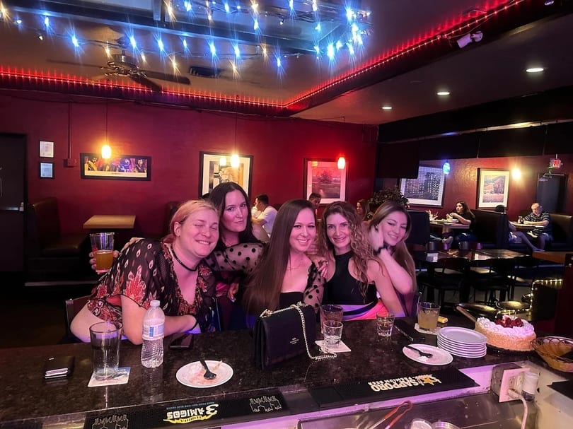 Five women smiling at a bar counter with drinks and a cake, in a dimly lit restaurant.